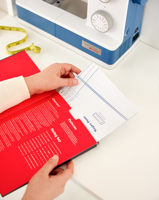 Person holding a red book with sewing patterns next to a sewing machine on a white table. This book is Ahead of the Curve by Jenny Rushmore.