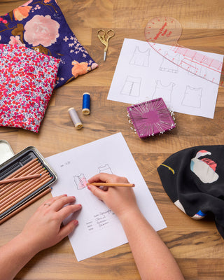 Person working on sewing or design project with fabric, patterns, and tools on a wooden table.