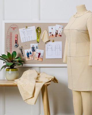 Tailor's workbench with dressmaking tools, fabric, and a mannequin wearing a beige dress. This is the Wyman Dress Sloper.
