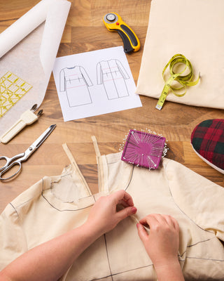Person sewing fabric with a pattern paper, scissors, and other tools on a wooden table. This is the Wyman Dress Sloper.