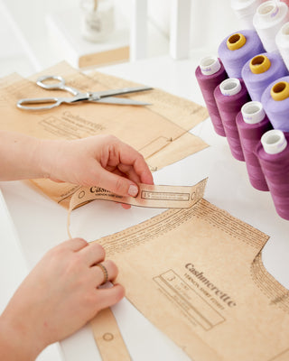 Hands holding Cashmerette sewing pattern pieces on a white desk, with purple thread and metal scissors nearby.