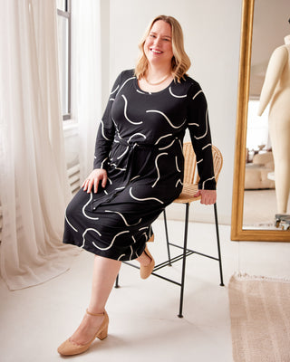 Woman sitting on a chair wearing a black dress with white patterns in a room. This is the Pembroke Dress sewing pattern from Cashmerette.