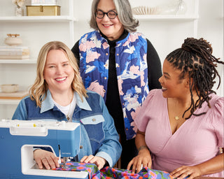 Three women working together at a sewing machine in a well-lit room.
