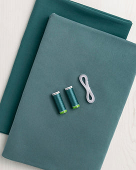 Two teal fabric cuts with spools of thread and a white elastic on a light gray background. This is the fabric kit for the Cashmerette Duxbury Dress sewing pattern.