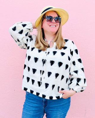 Person wearing a white blouse with black abstract patterns, blue jeans, and a yellow hat against a pink background. This top is the Alton Blouse sewing pattern from Cashmerette.