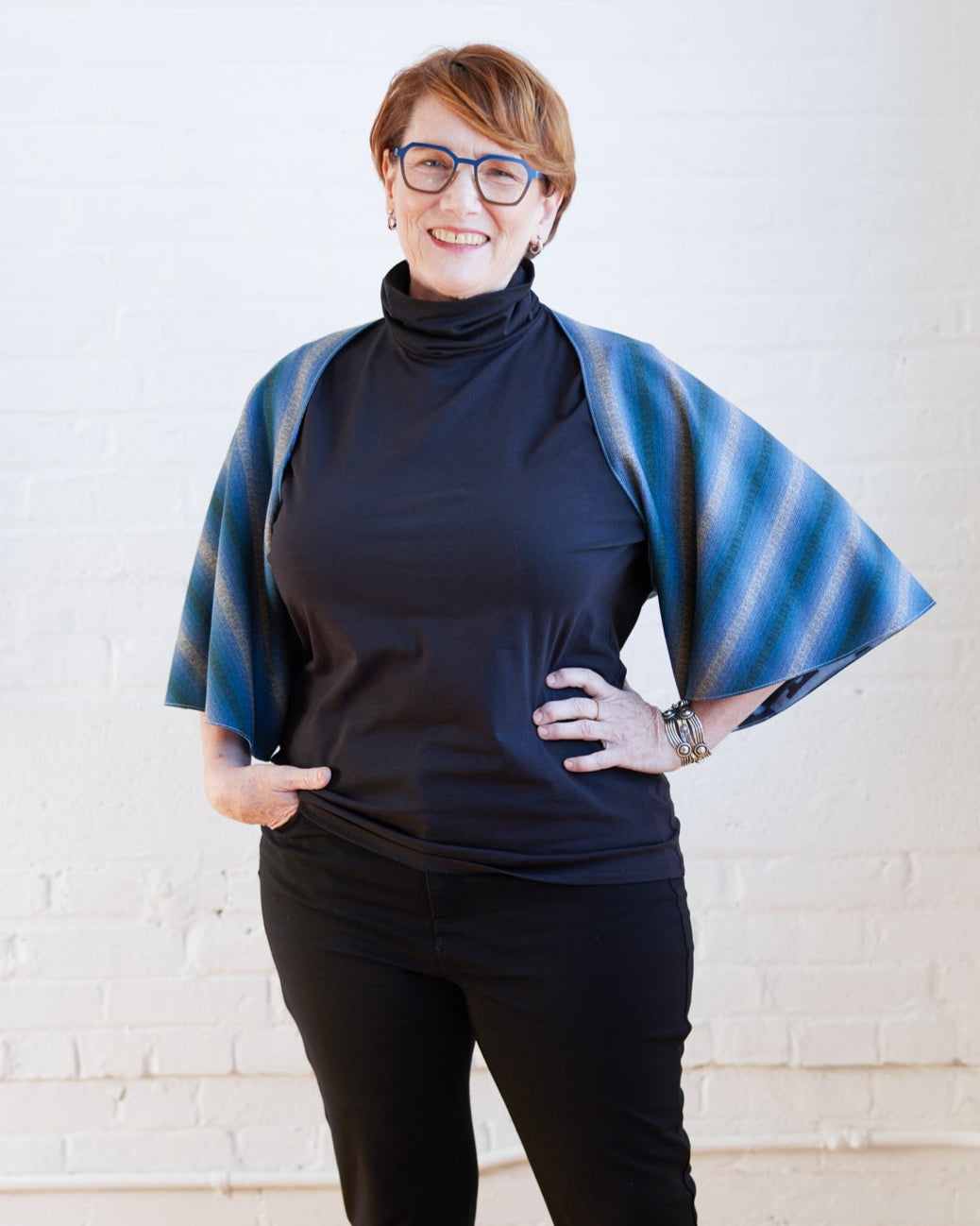 Person wearing a blue and green striped poncho over a black outfit against a white brick wall. The topper is the Caroll Shrug sewing pattern from Cashmerette.