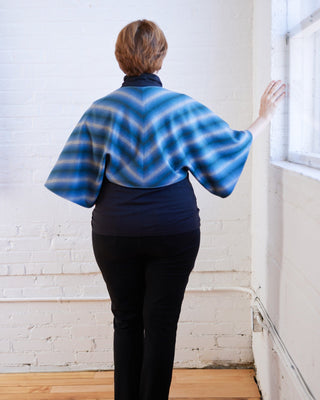 Person wearing a blue and gray patterned shawl standing in a room with white walls and wooden floor. The topper is the Caroll Shrug sewing pattern from Cashmerette.
